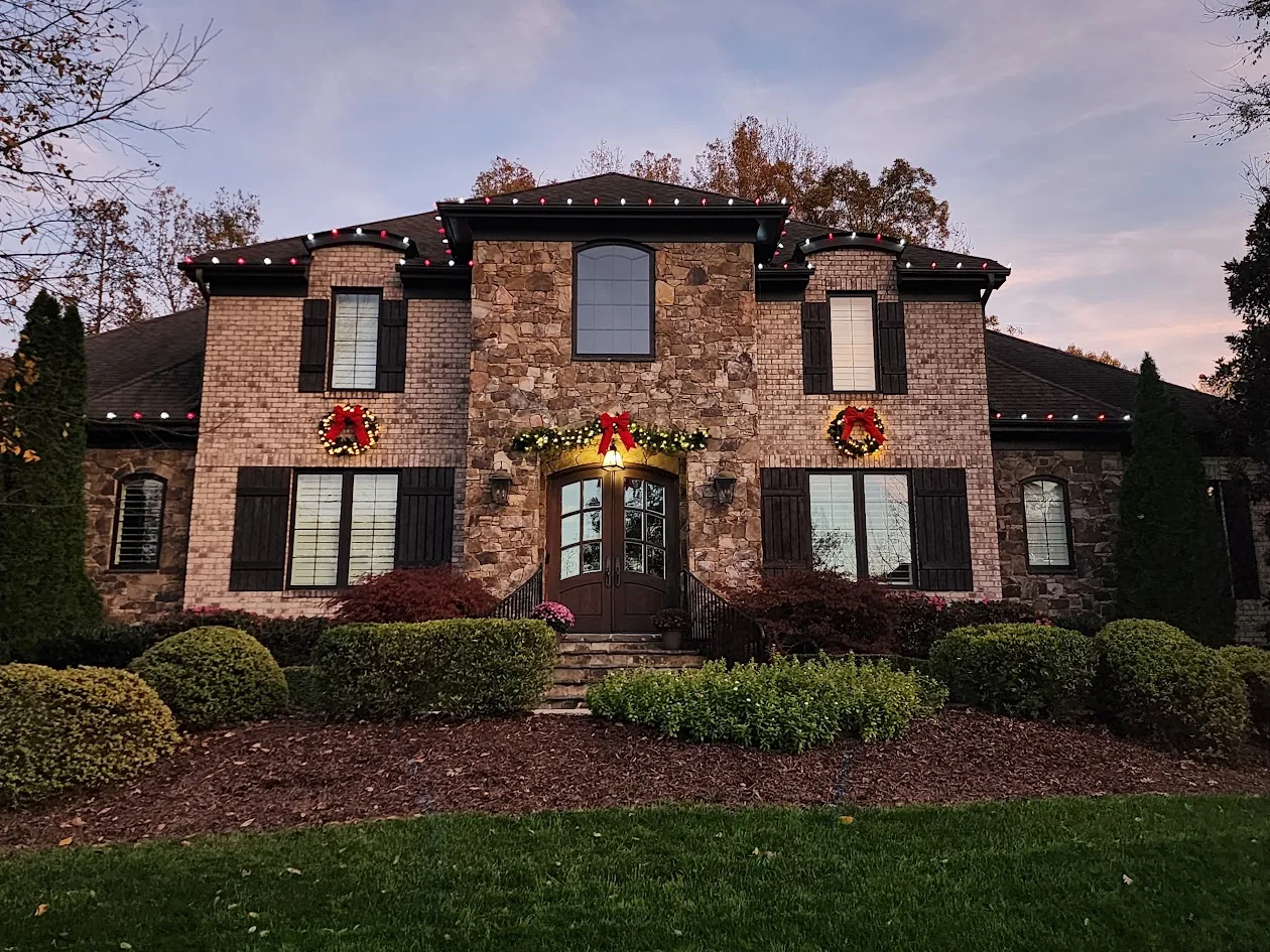 Red and warm white roofline lighting, wreaths, and entry greenery on a large stone-and-brick home