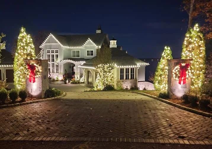 Warm-white roofline with two lit evergreens and red bows along a paver drive
